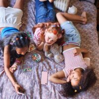 children lying on a bed, using their phones and eating junk food