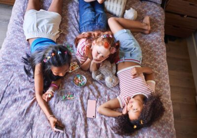 children lying on a bed, using their phones and eating junk food