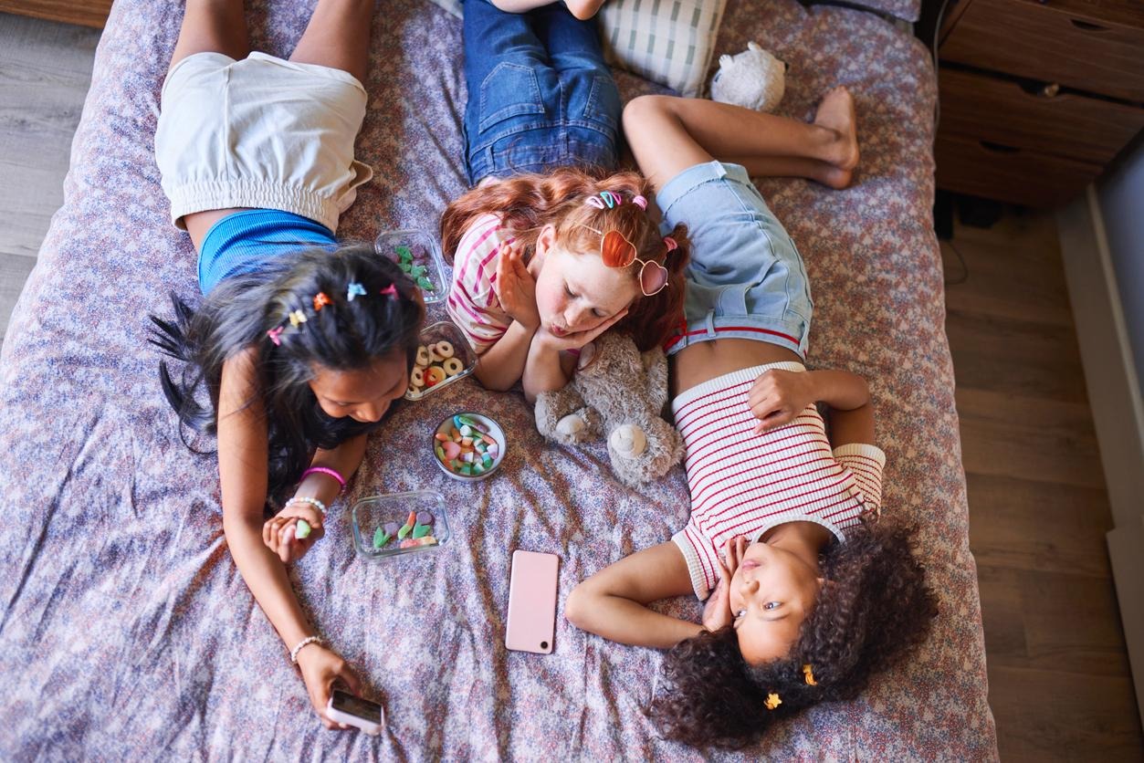 children lying on a bed, using their phones and eating junk food