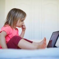 A little girl sitting alone on a bed, looking at a tablet