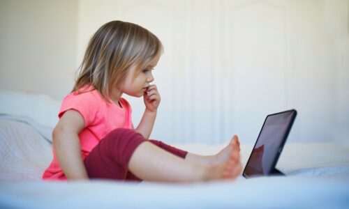A little girl sitting alone on a bed, looking at a tablet