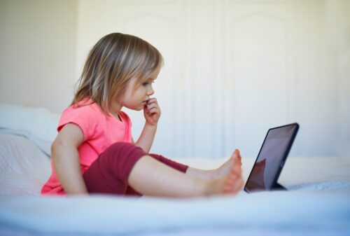 A little girl sitting alone on a bed, looking at a tablet
