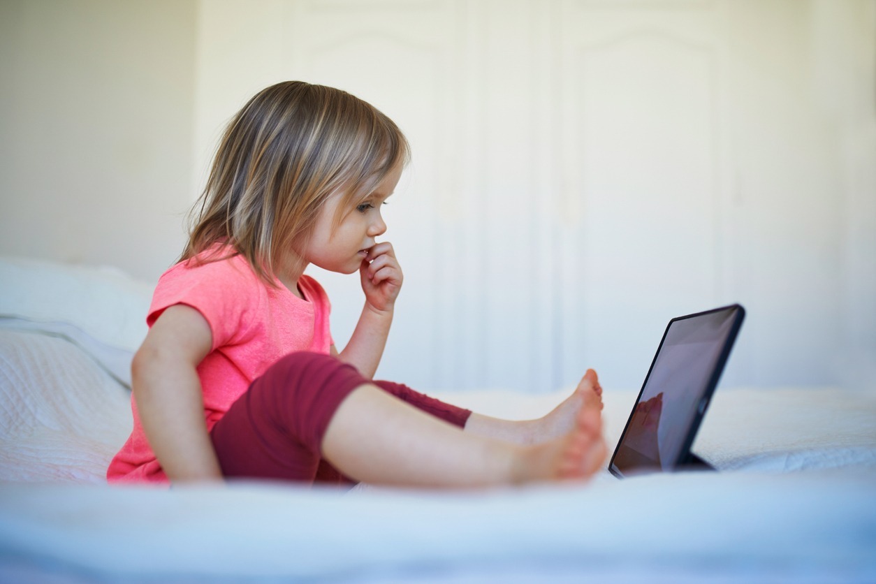 A little girl sitting alone on a bed, looking at a tablet